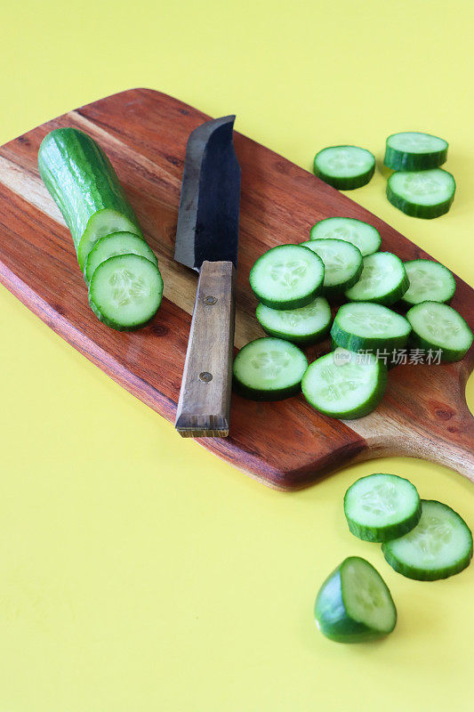 黄瓜(Cucumis sativus)和菜刀在木制砧板上的图像，整个水果和切片显示绿色的皮，白色的肉和白色的种子，黄色背景，高架视图，重点在前景